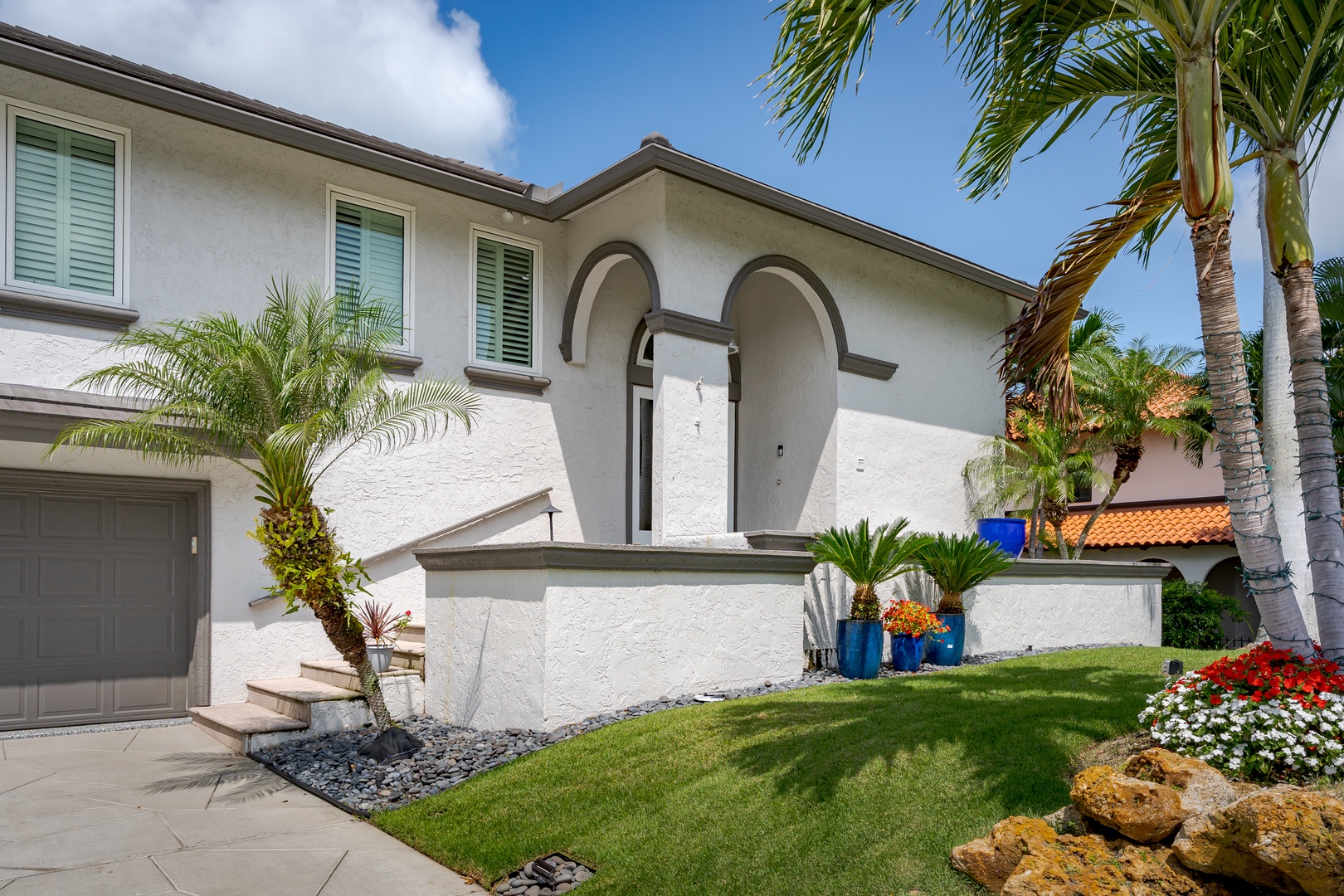 A Mediterranean-style vacation home with arched entry and lush tropical landscaping welcomes guests with vibrant gardens and swaying palms under sunny skies.