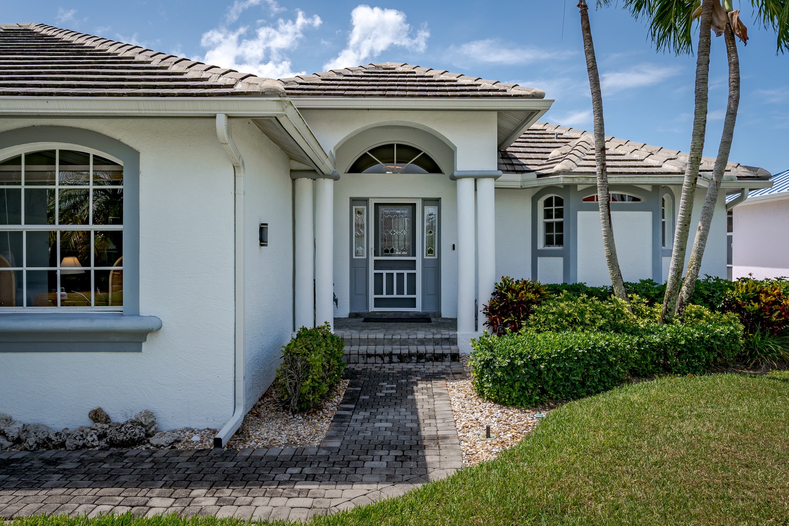 Elegant single-story home featuring a columned entrance, tiled roof, and manicured landscaping with swaying palm trees in a peaceful residential setting.