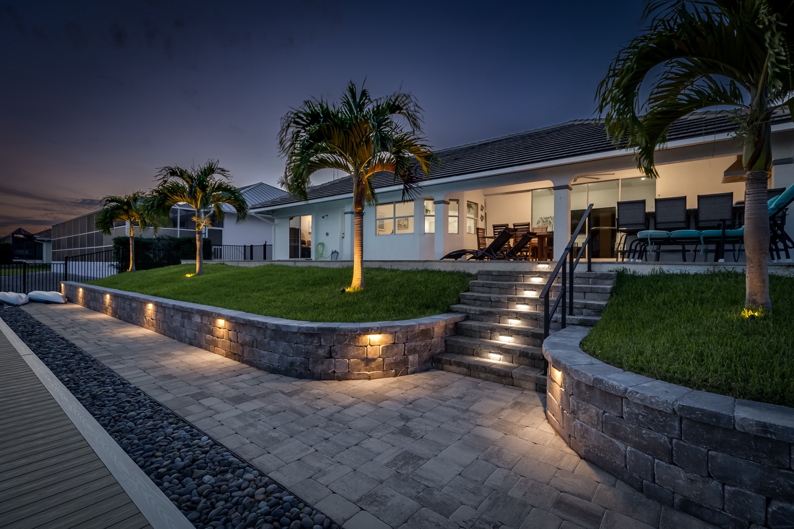 A modern property exterior features illuminated landscaping and terraced outdoor areas in a peaceful neighborhood setting at dusk.