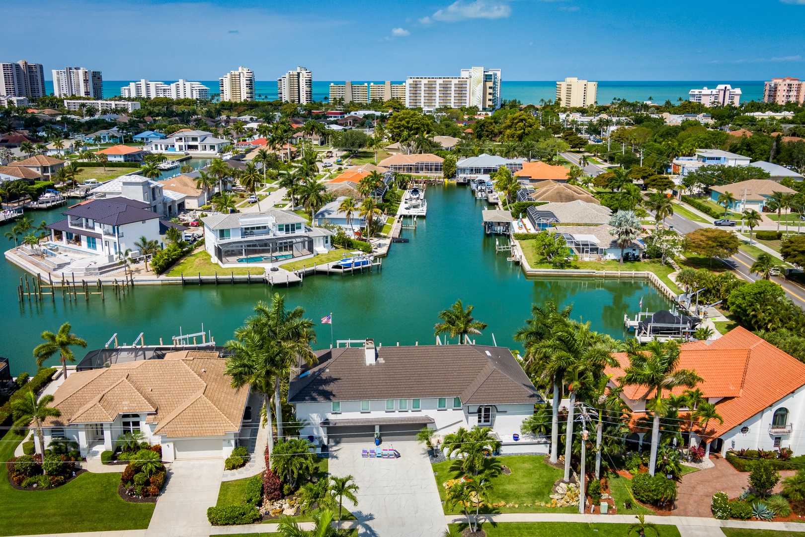 Aerial view of a waterfront community featuring canal-front homes with private boat docks, lush palm trees, and Gulf Coast high-rises visible beyond the turquoise waters.