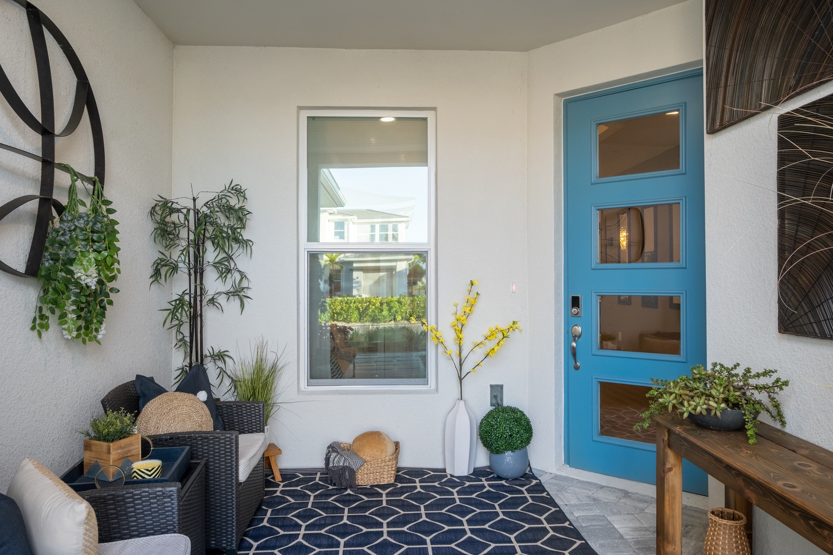 Relax on this charming covered porch with wicker seating, vibrant blue door, and lush greenery creating your perfect welcome retreat.