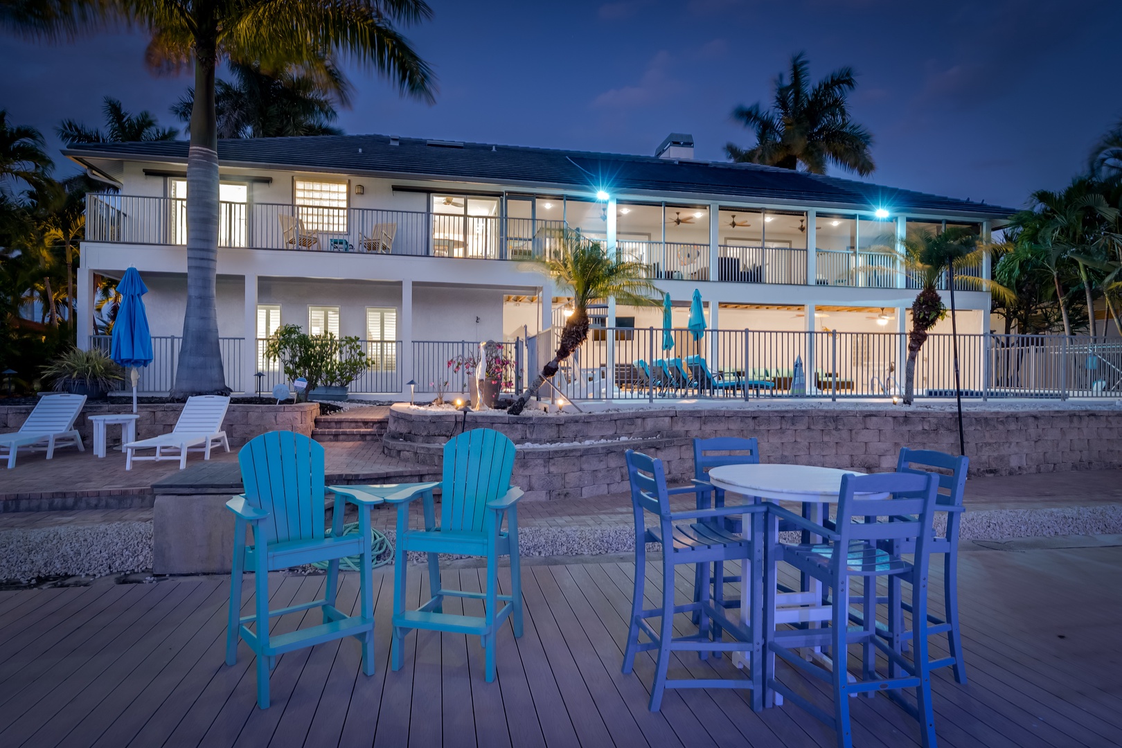 A waterfront property framed by tropical palms, featuring a spacious deck with outdoor dining and lounge areas overlooking the water—an ideal setting for gathering at dusk.