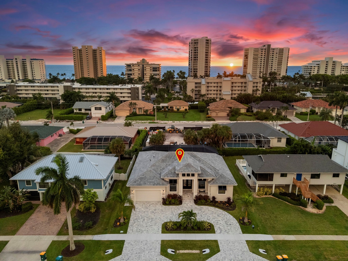 An aerial view captures the property nestled in a peaceful neighborhood, with palm-lined streets and a stunning sunset painting the sky in vibrant hues over the distant cityscape.