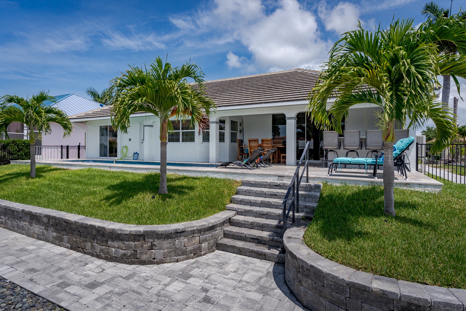 Modern tropical property featuring expansive covered patio with lounge seating, manicured lawns framed by palm trees, and private pool area—designed for relaxation in a sunny coastal setting.