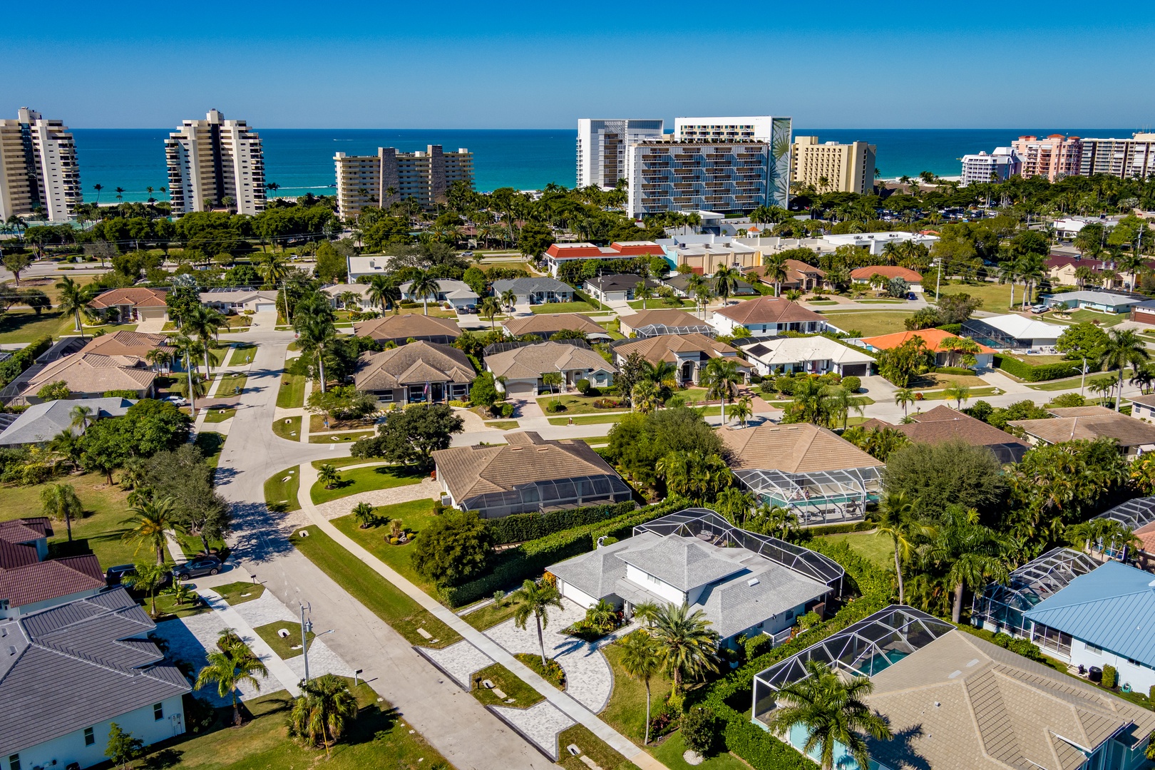 Aerial view of a coastal residential neighborhood with palm-lined streets, private pools, and nearby beachfront high-rises.