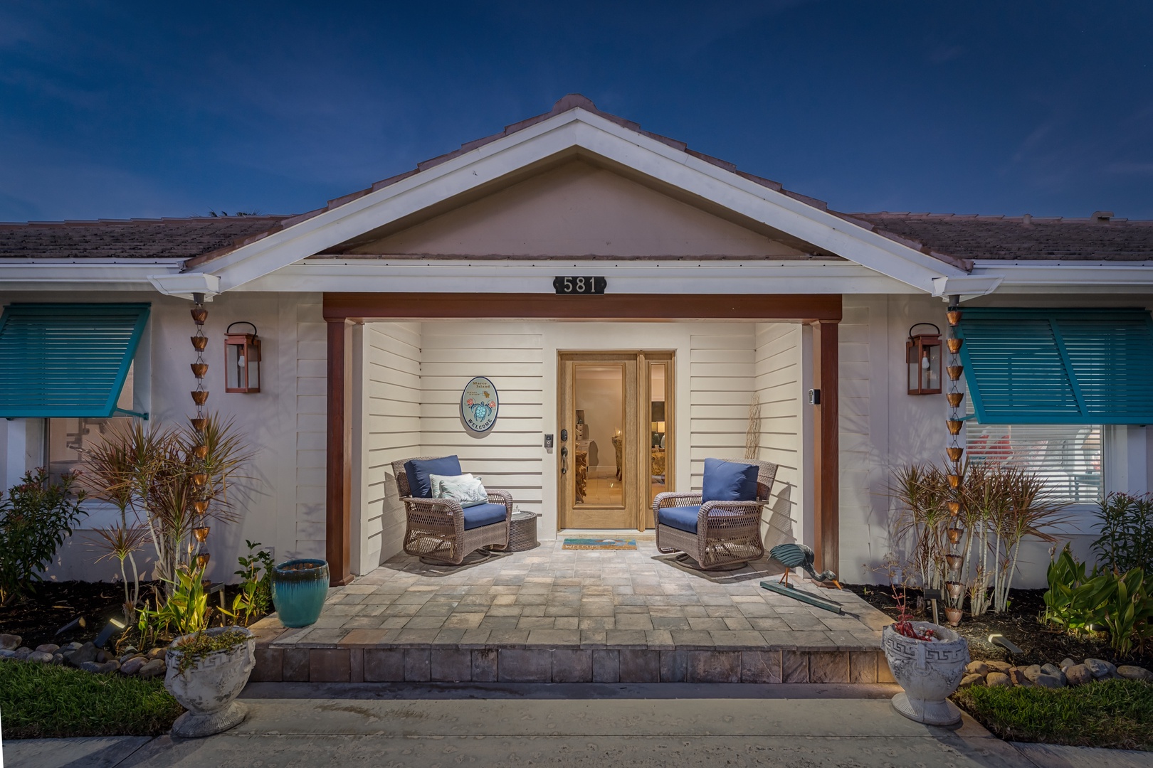 Charming coastal home with welcoming front porch featuring comfortable seating and tropical plants under a deep blue evening sky.