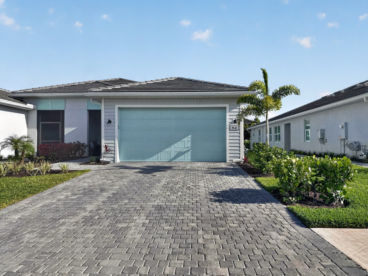 Modern single-family home with two-car garage and tropical landscaping in a residential neighborhood. The property features paver driveway and easy parking access.