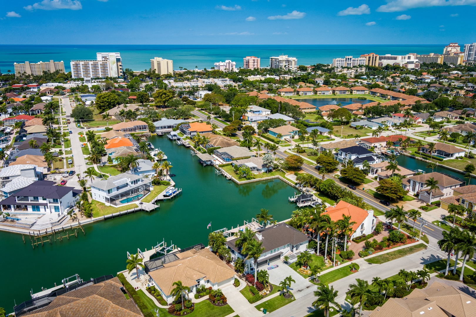 Aerial perspective of a coastal neighborhood featuring waterfront homes with private docks, tree-lined canals, and nearby beach access, located in a vibrant resort area.