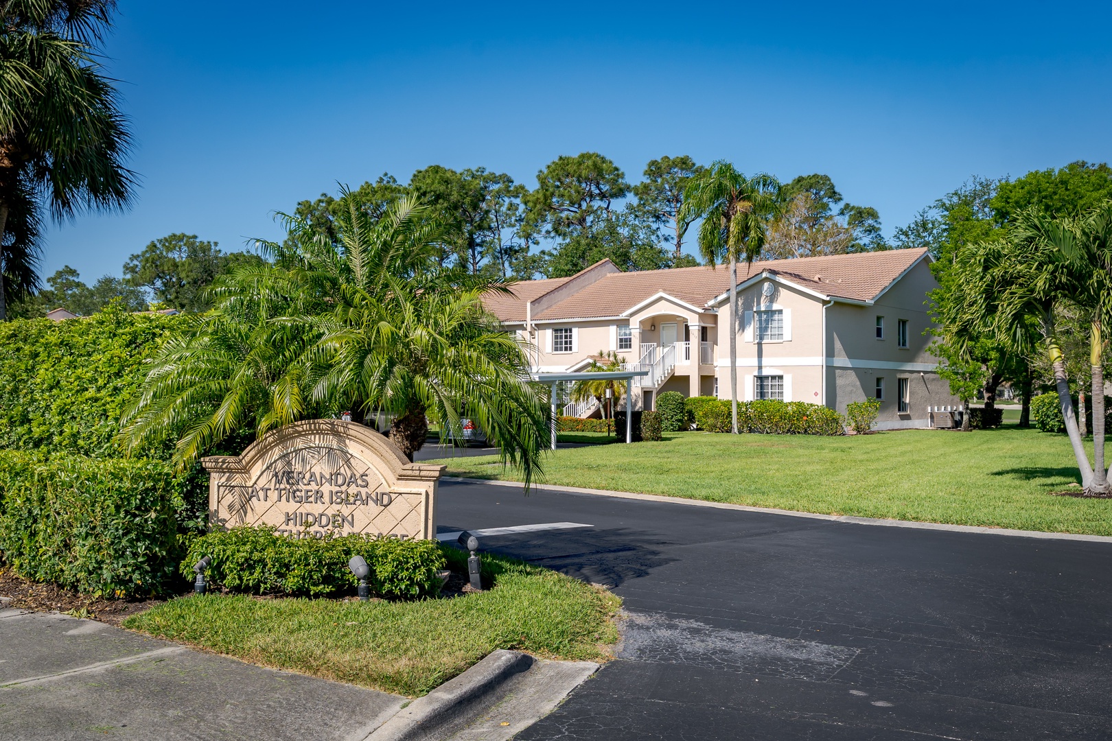 The property entrance features lush tropical landscaping with palm trees and manicured lawns, located within a peaceful residential community surrounded by mature greenery.