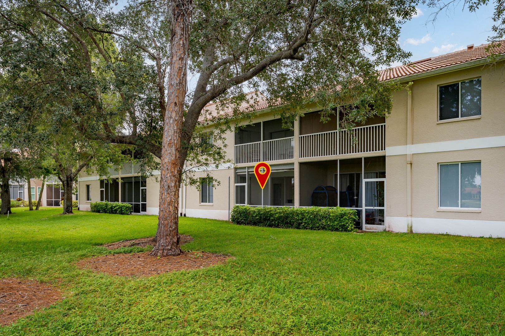 The property features a beige two-story building with tiled roofing, set within a lush green lawn area surrounded by mature trees. Covered balconies and outdoor spaces provide a peaceful setting for guests arriving at this tranquil retreat.