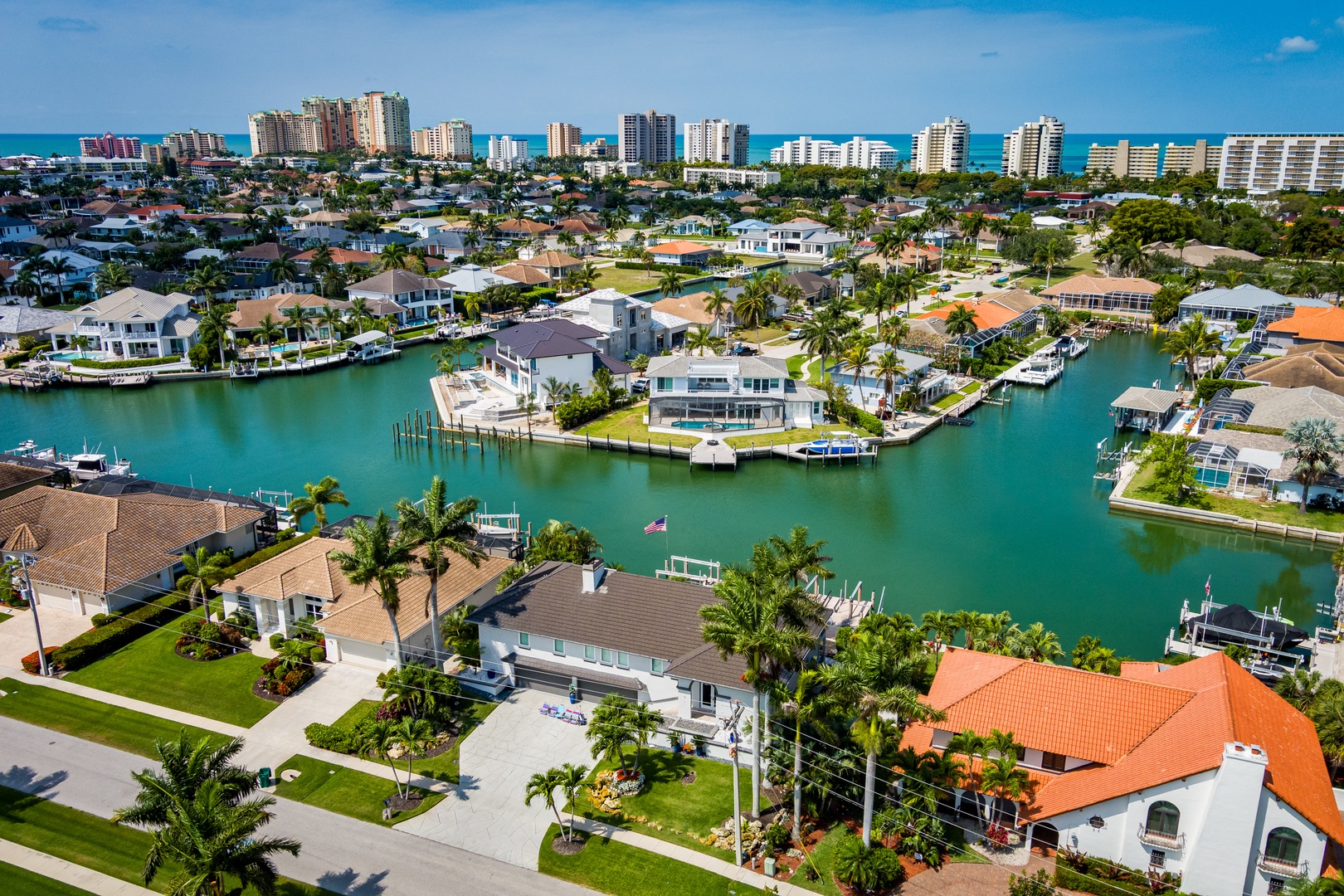 Aerial view of a prestigious waterfront community with luxury homes lining turquoise canals, private docks, and the Gulf of Mexico coastline in the distance.