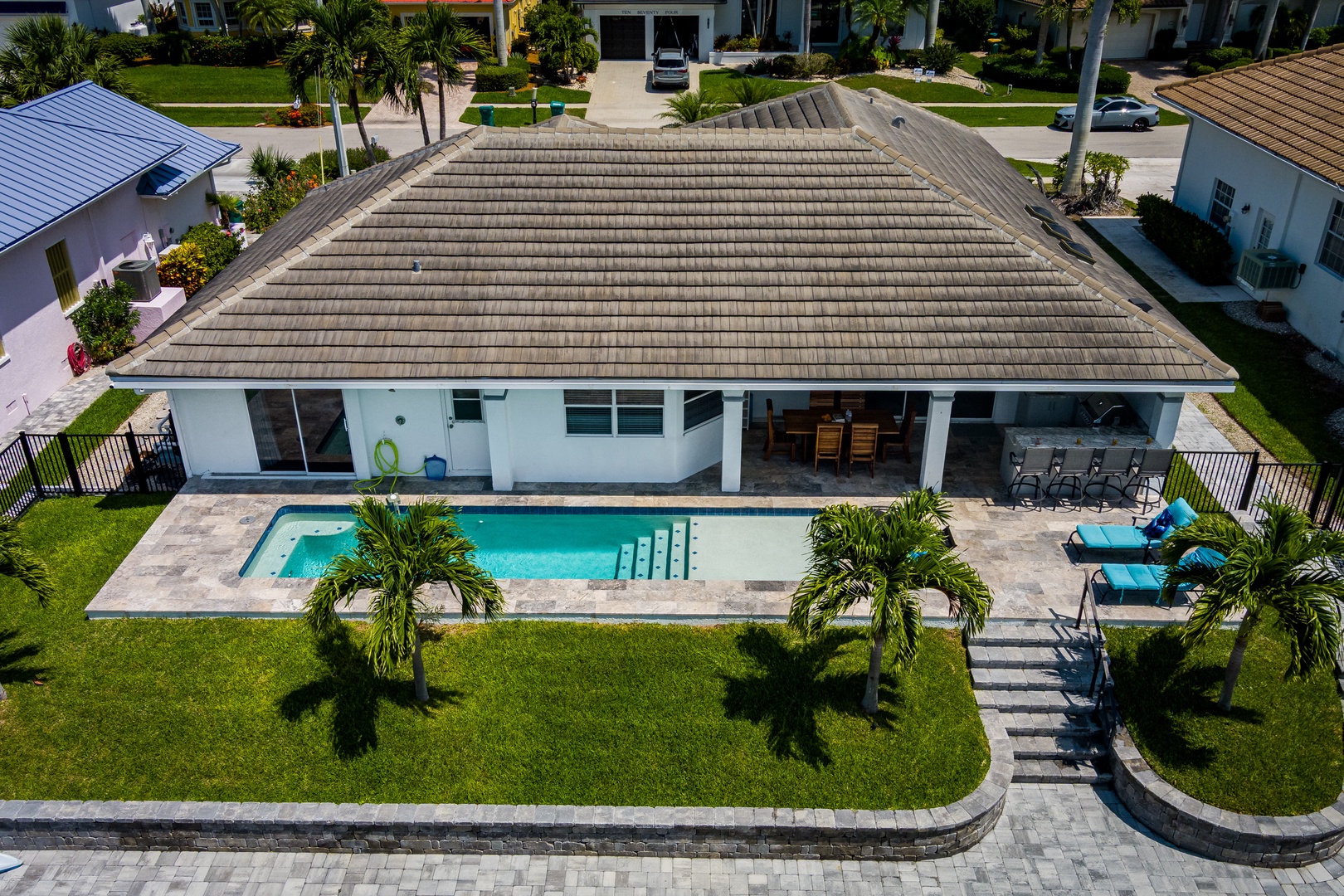 Aerial view of a modern single-story vacation home featuring a private pool, spa, and covered patio surrounded by tropical landscaping in a quiet residential neighborhood.