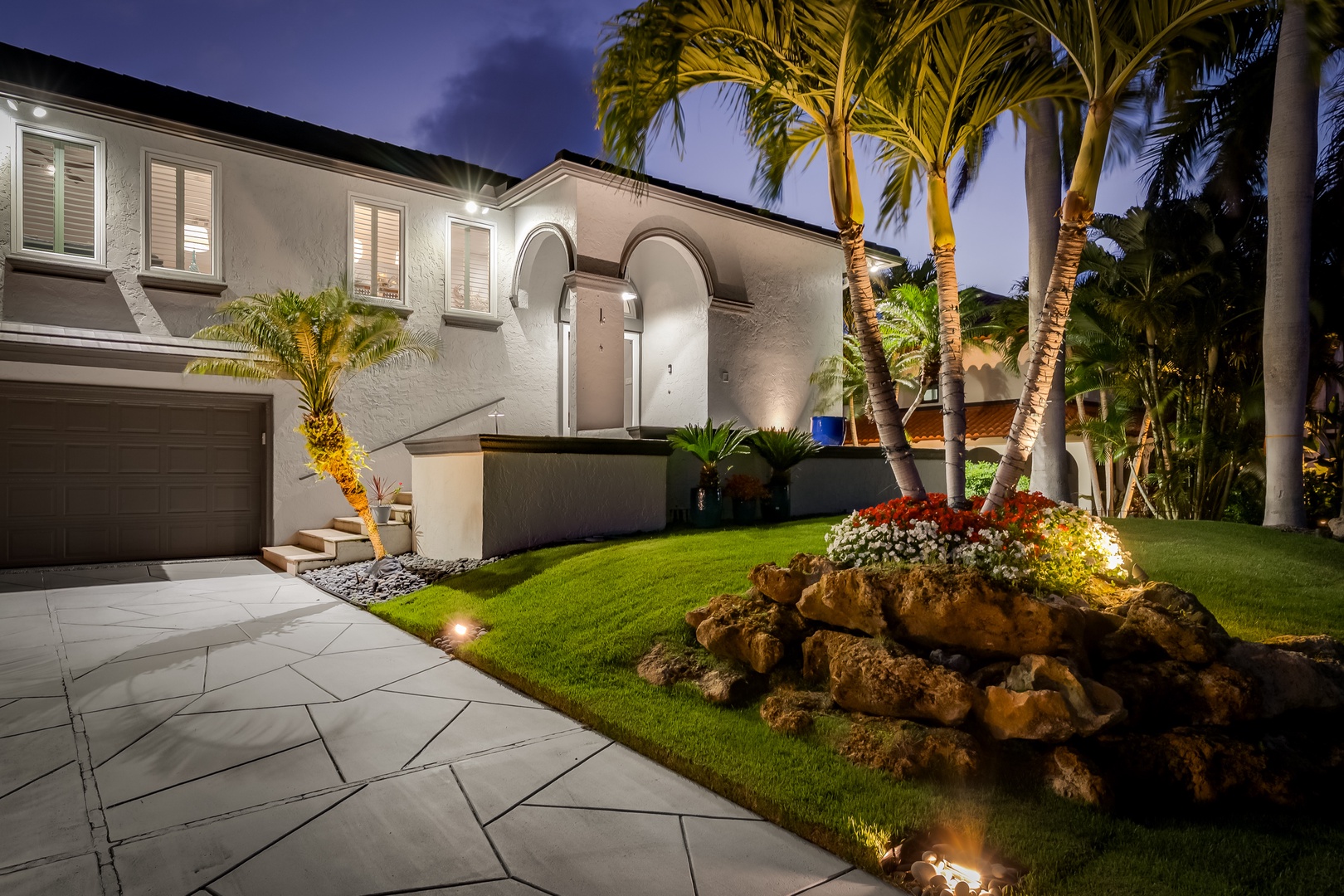 This elegant property features a Mediterranean-inspired facade with arched entryway, lush tropical landscaping with towering palms, and beautifully illuminated grounds—a stunning retreat captured at twilight.