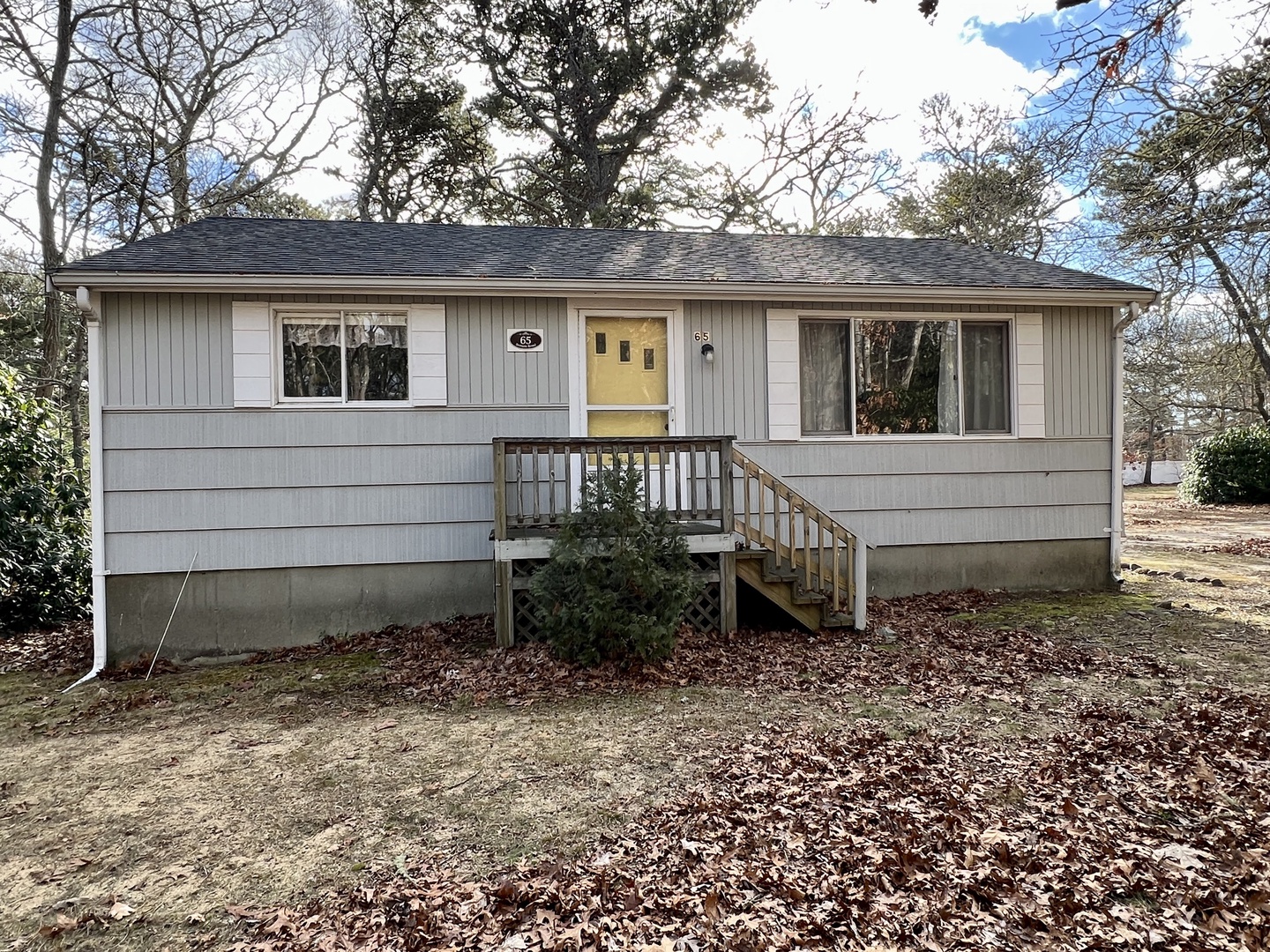 A charming single-story home featuring a welcoming yellow front door and wooden steps, nestled among mature trees in a peaceful wooded setting.