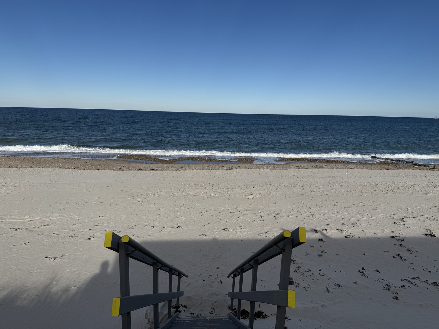 Pristine sandy beach stretches beneath clear blue skies, with gentle waves rolling onto shore and beach access stairs leading to the water.