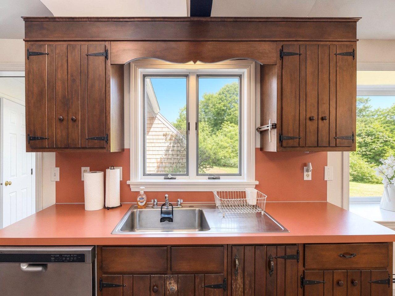 Your charming kitchen features warm wood cabinetry and vibrant countertops, perfect for preparing morning coffee while overlooking lush summer greenery through the window.