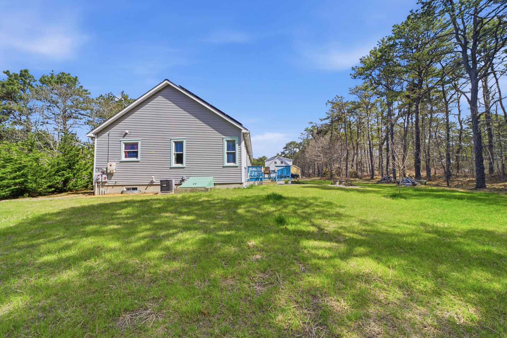 Charming gray house sits peacefully on spacious green lawn, surrounded by mature trees and natural woodland setting.