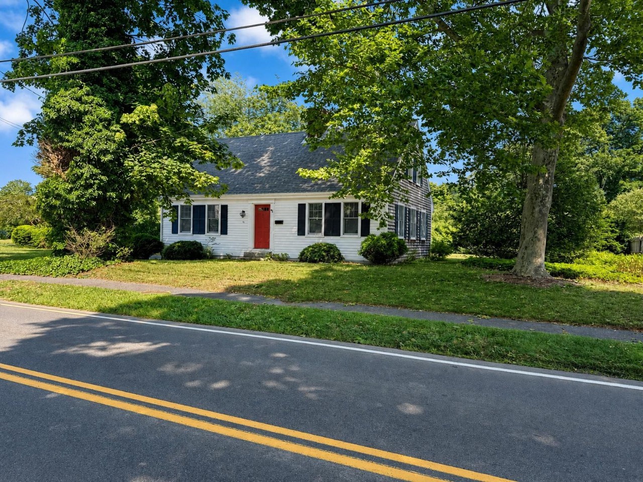 Classic white Cape Cod-style cottage with charming red door and black shutters, nestled among mature trees on a peaceful residential street.