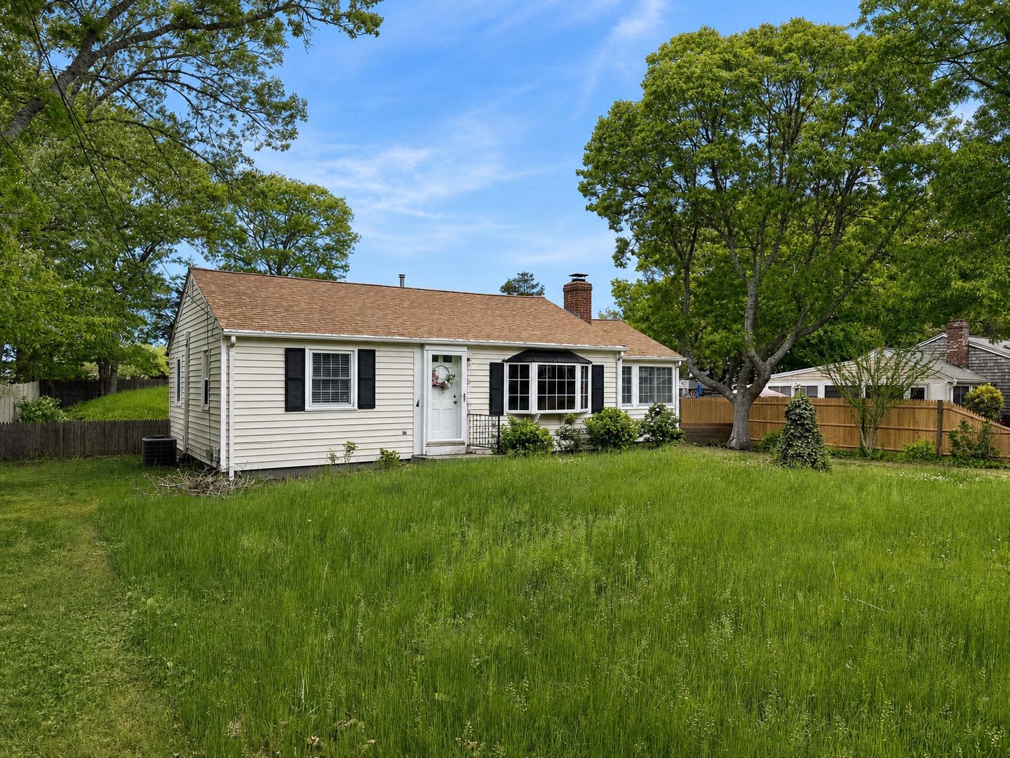 A charming single-story cottage with classic white siding and black shutters, surrounded by mature trees and a spacious lawn offering a peaceful suburban setting.