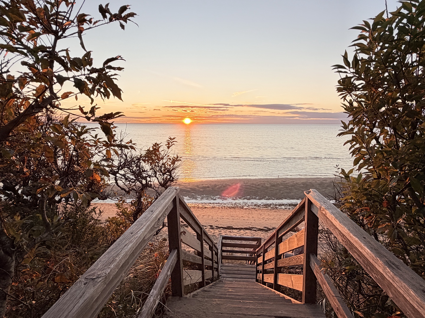 Wooden boardwalk leads through coastal vegetation to a pristine beach where golden sunset paints the horizon over calm waters.