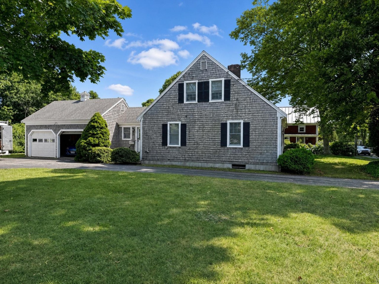 Classic Cape-style home with charming weathered shingles, mature trees, and attached garage in a peaceful residential neighborhood.