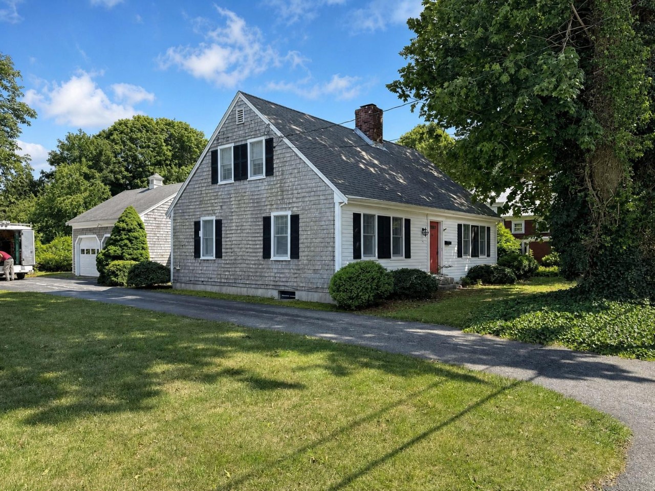 Classic Cape Cod home with weathered shingle siding, mature landscaping, and private driveway parking set on a spacious green lawn surrounded by tall shade trees.