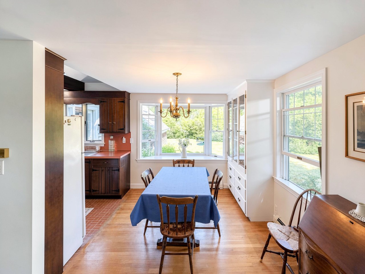 Gather around the dining table in this bright, welcoming space where natural light pours through large windows framing lush garden views. The warm wood cabinetry and classic chandelier create a homey atmosphere perfect for shared meals.