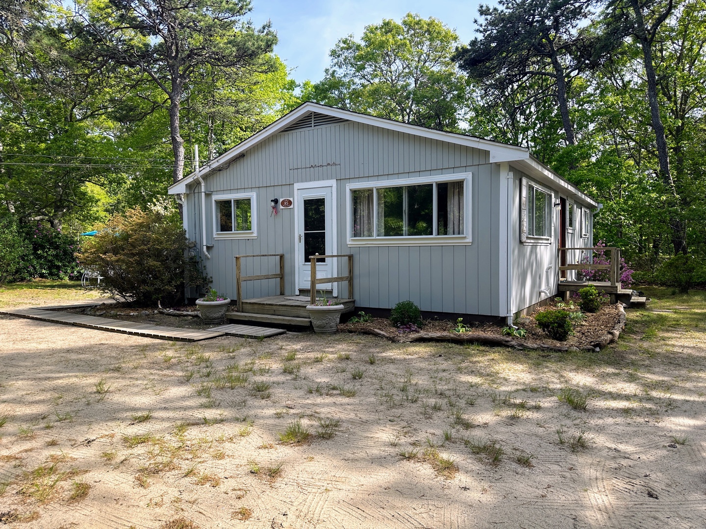 Charming gray cottage nestled among mature trees offers a peaceful woodland retreat with welcoming front porch and natural landscaping.