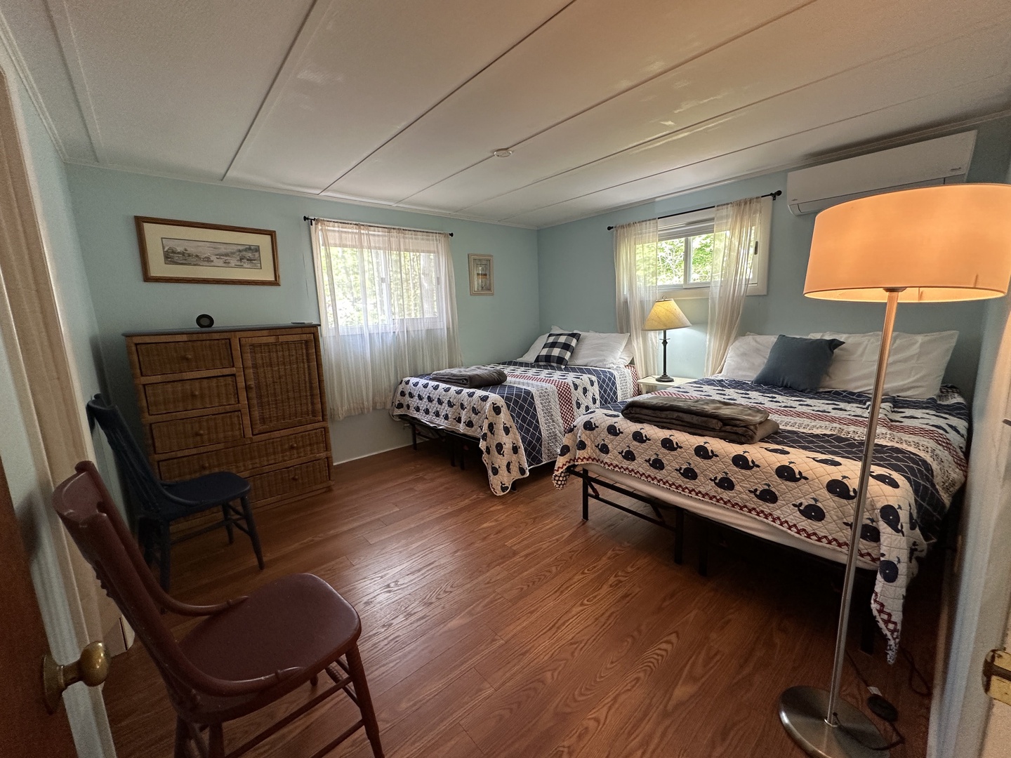 Your comfortable twin bedroom features charming coastal décor, warm wood floors, and plenty of natural light filtering through soft curtains.