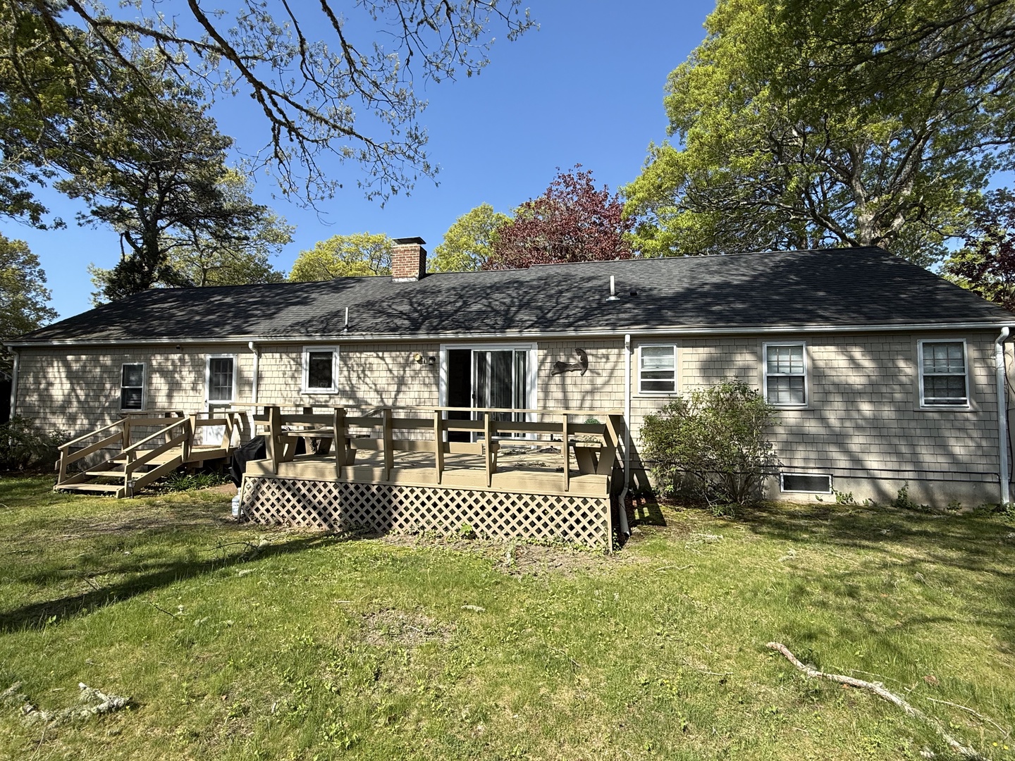A charming single-story vacation home features a spacious wooden deck with outdoor seating, perfect for relaxing under the shade of mature trees in this peaceful, tree-lined setting.