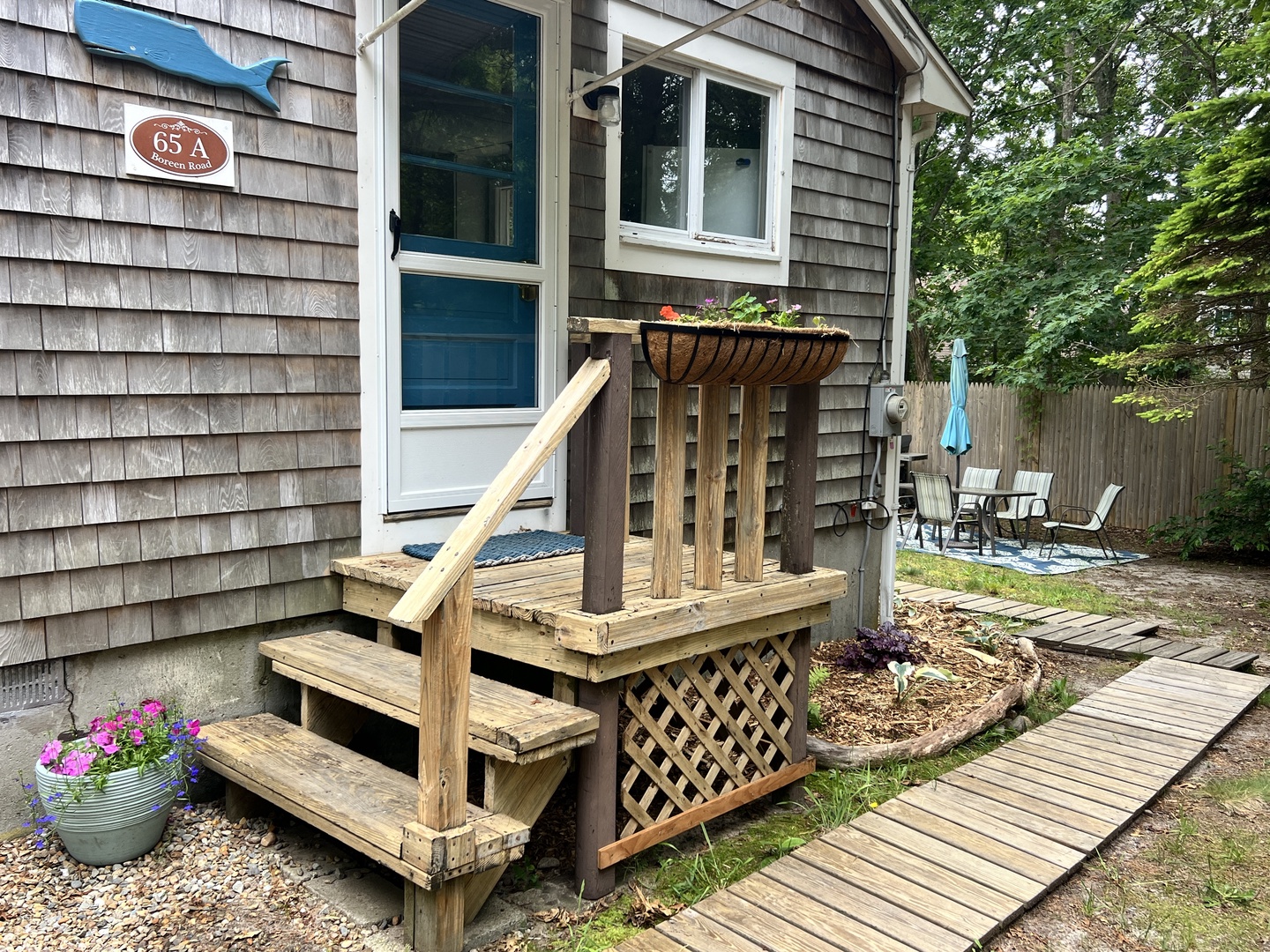 Charming cedar-shingled cottage entrance welcomes guests with wooden steps, flower baskets, and private patio beyond.