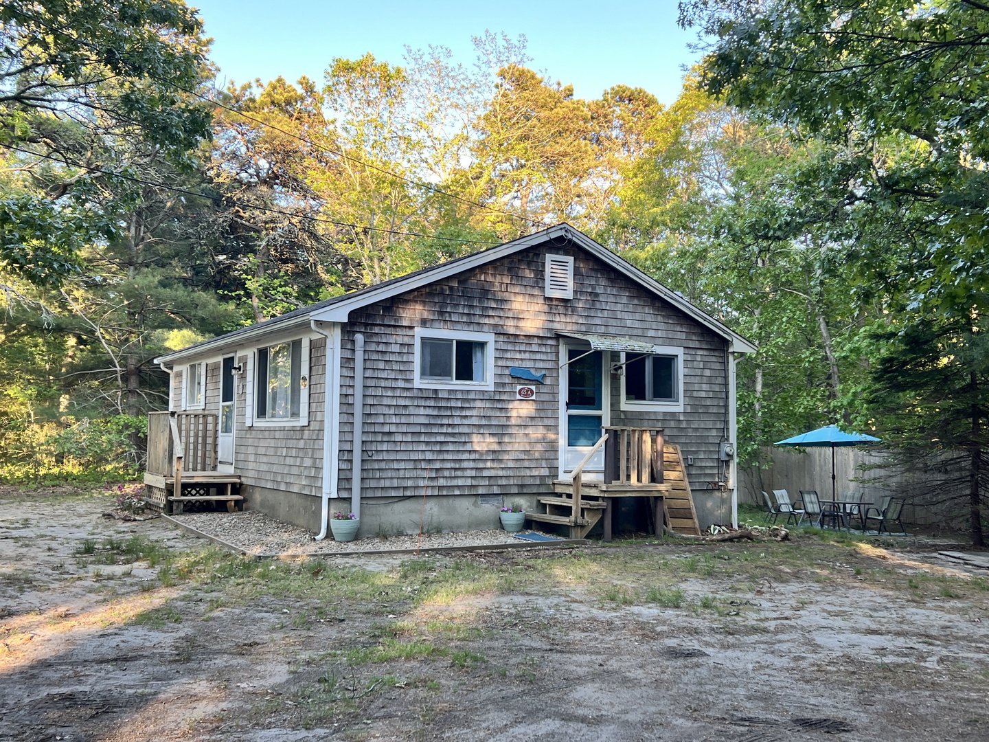 Charming shingled cottage nestled among mature trees with outdoor seating area for peaceful woodland relaxation.