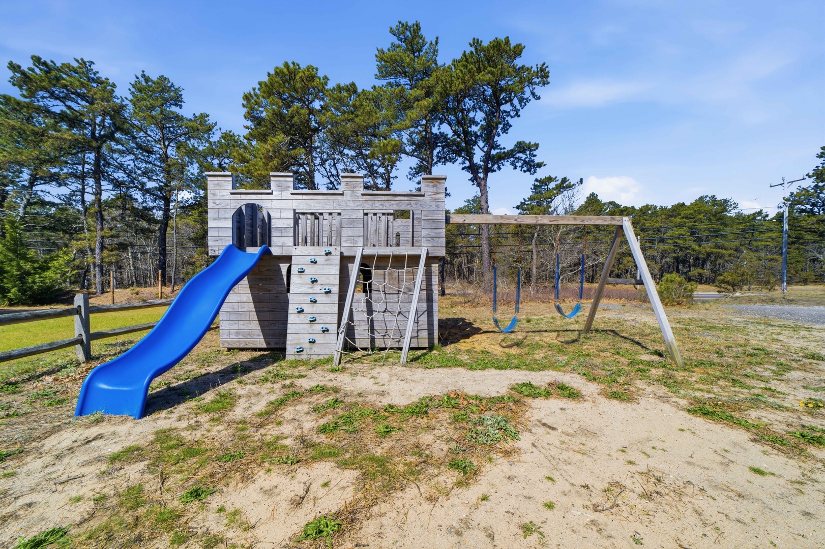 A castle-themed playground with slide, climbing walls, and swings surrounded by peaceful wooded landscape.