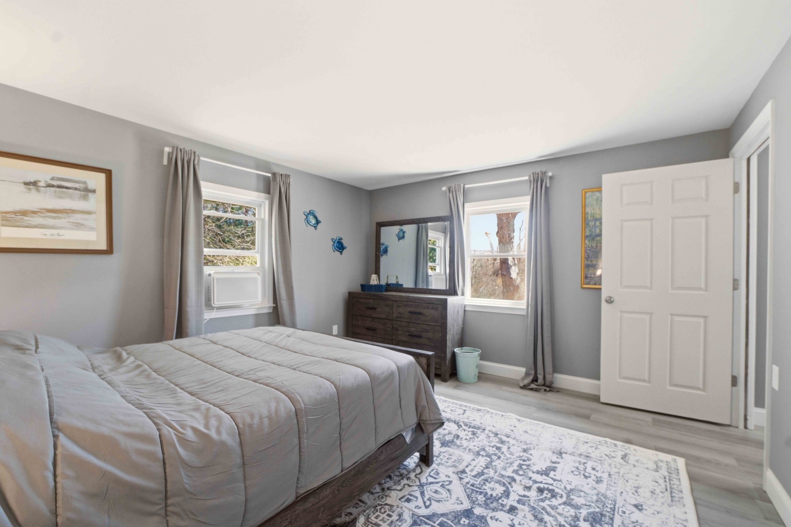 Rest peacefully in this serene bedroom featuring soft gray walls, comfortable bedding, and bright natural light from dual windows.