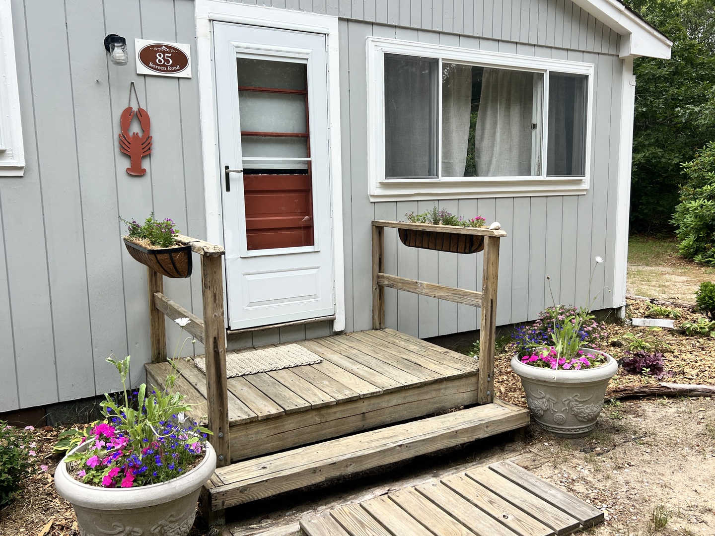 Charming cottage entrance with wooden deck and colorful flower displays creates a welcoming arrival to your coastal getaway retreat.