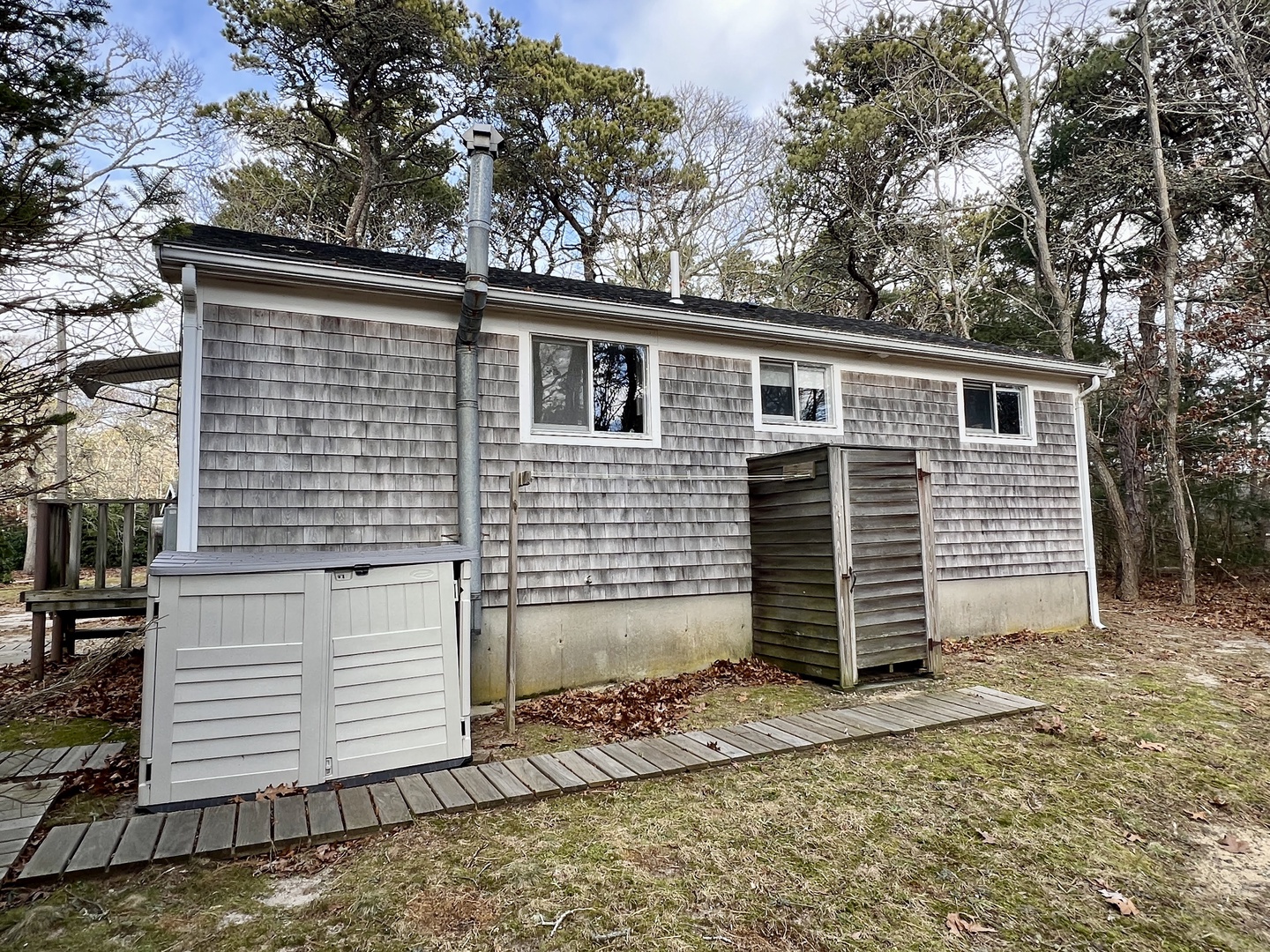 A charming cedar-shingled cottage nestled among mature trees, featuring classic New England architecture with an outdoor shower for beach days.