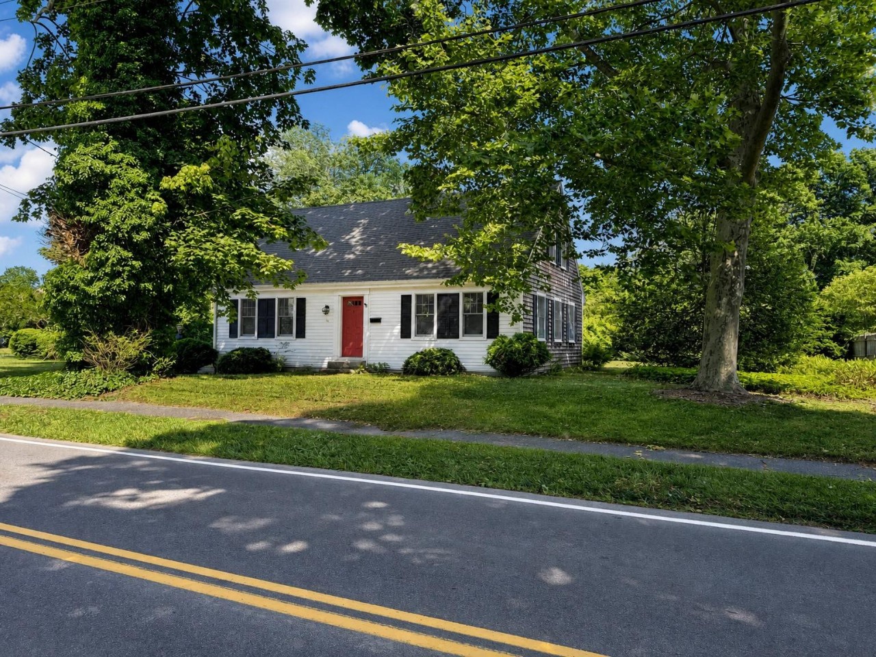 A charming Cape Cod-style cottage with a welcoming red door sits nestled among mature shade trees on a peaceful residential street.