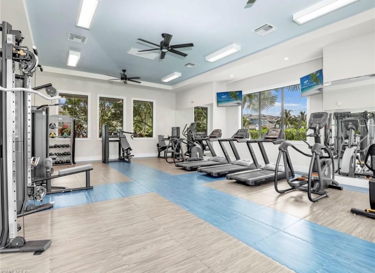 Step into your fully-equipped fitness center with modern cardio and strength equipment, bright natural light, and tropical garden views through large windows.