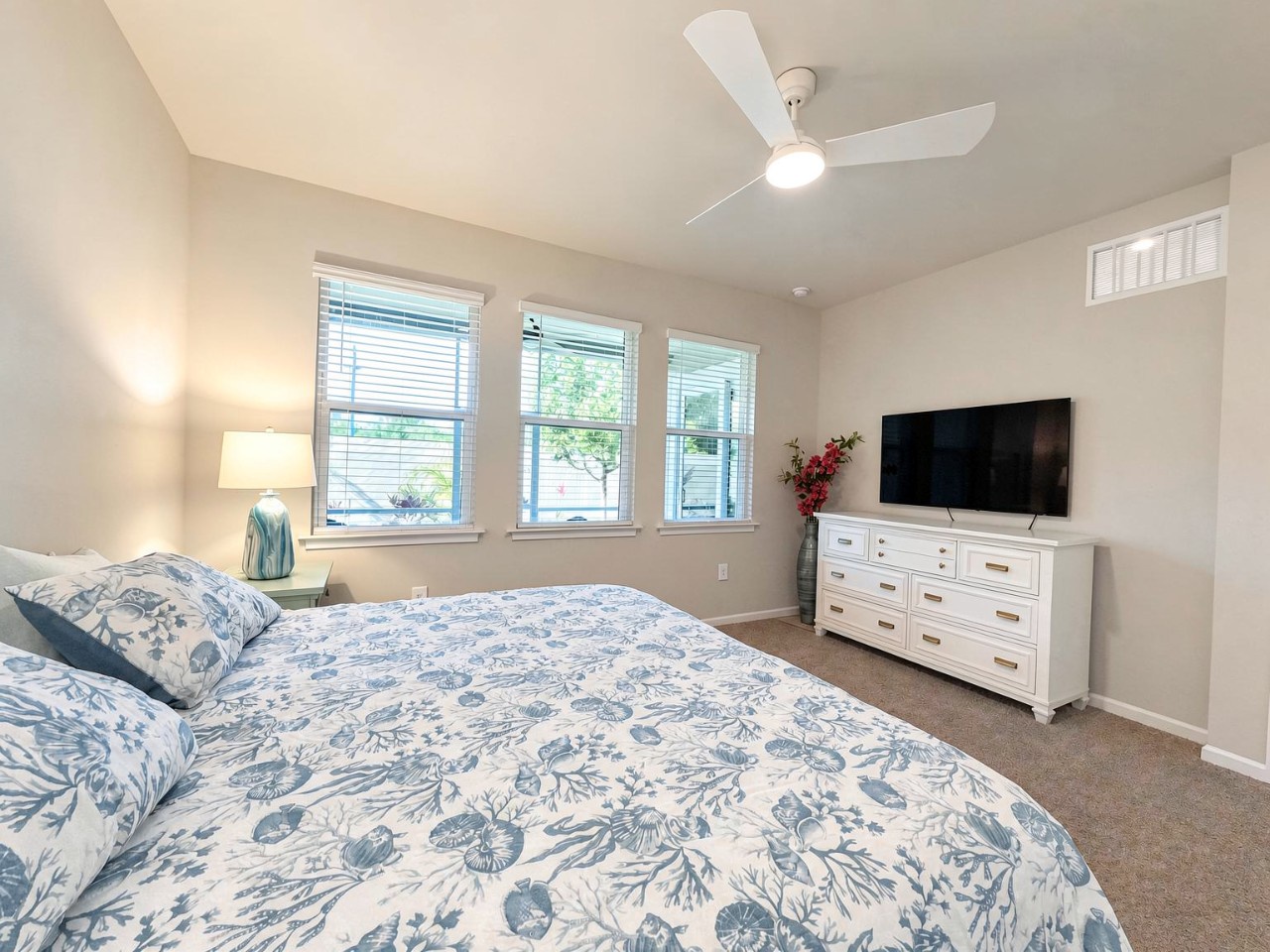 Your serene bedroom retreat awaits, with a cozy bed dressed in soft linens, a ceiling fan keeping you cool, and natural light streaming through multiple windows for a bright, airy feel.