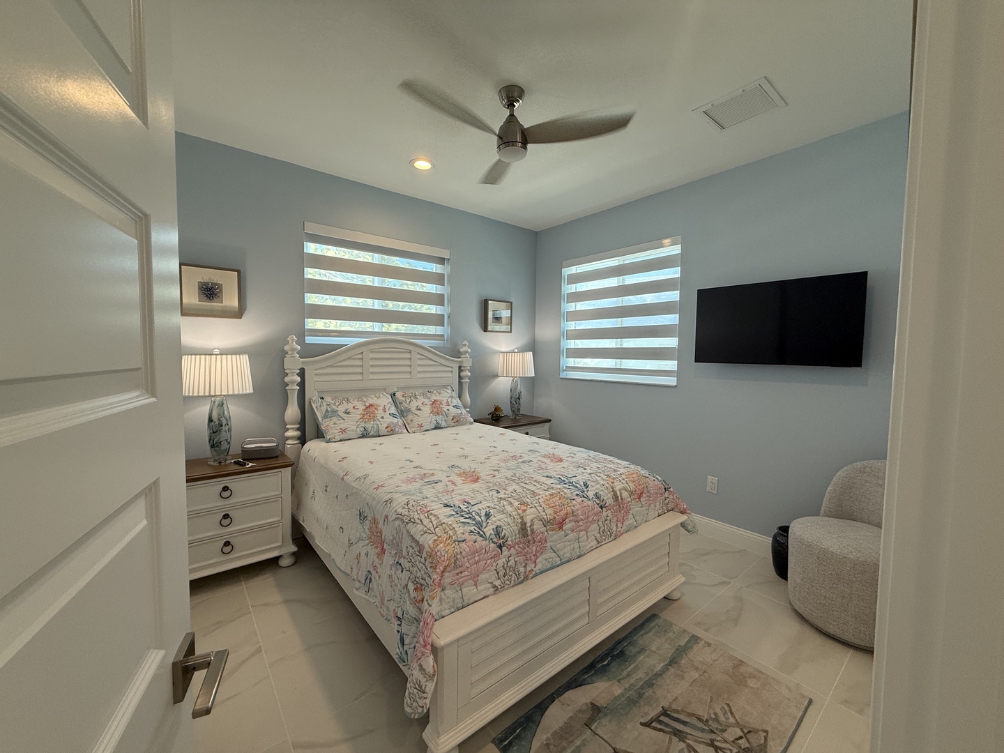 Drift off in your serene bedroom featuring soft blue walls, a ceiling fan, and botanical bedding that whispers coastal comfort.
