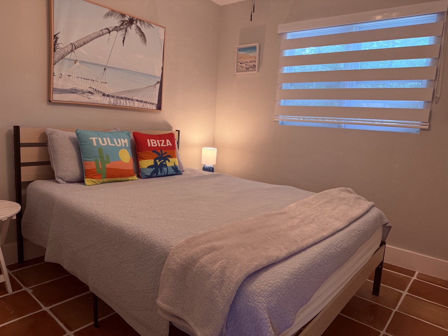 Your comfortable bed awaits beneath warm wooden beams, with crisp white linens and colorful beach towels ready for your stay. A cozy window lets in natural light to brighten your mornings.