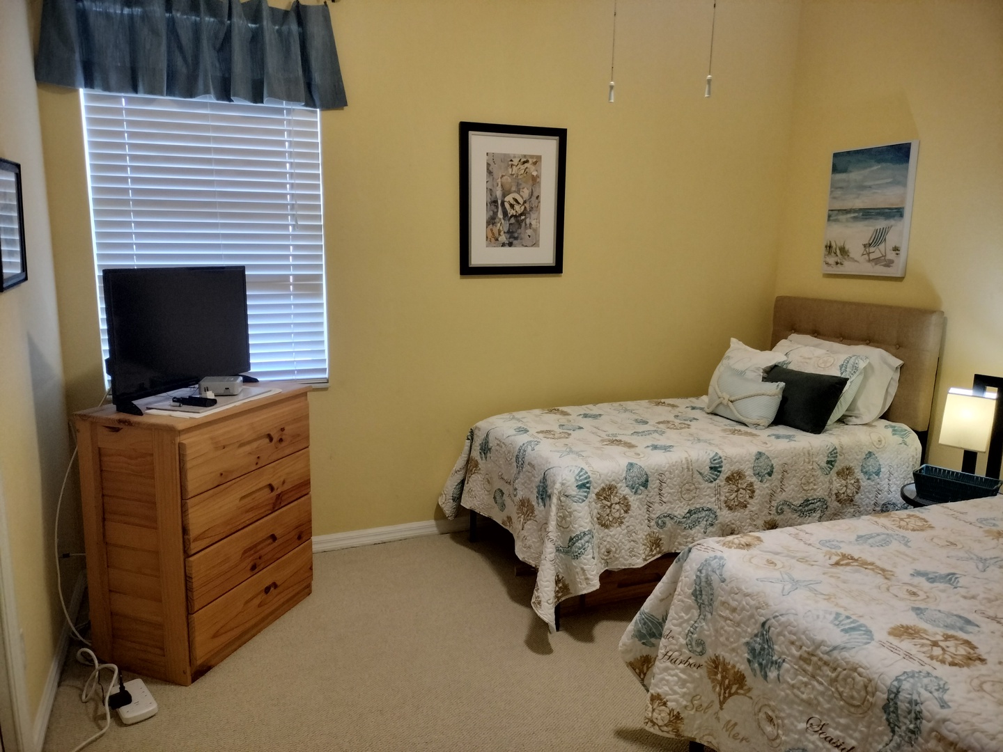 Unwind in your cozy guest room with new twin beds, warm yellow walls, and beach-themed art that sets a peaceful coastal mood for your stay.