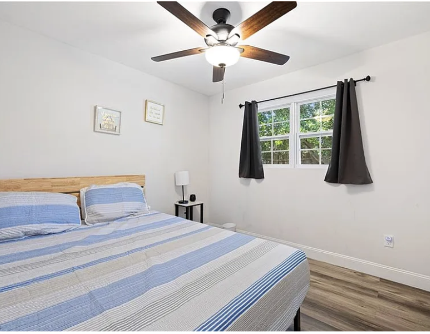 Your bright bedroom features a cozy bed with fresh linens, a ceiling fan for comfortable sleep, and a charming window framing views of lush greenery—your peaceful retreat awaits.