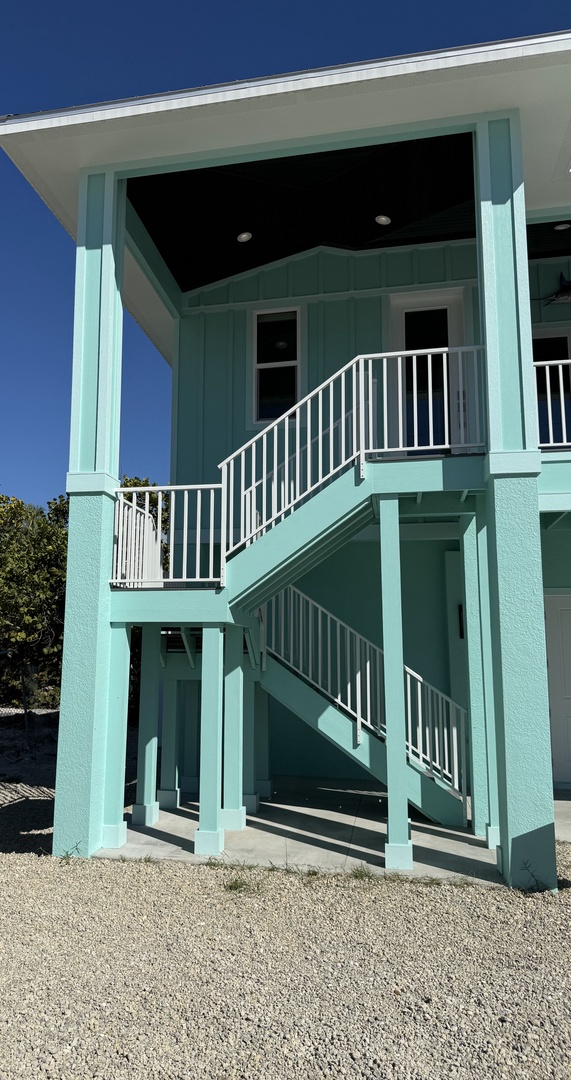 Charming turquoise beach house with white stairs leading to covered balcony under clear blue skies.