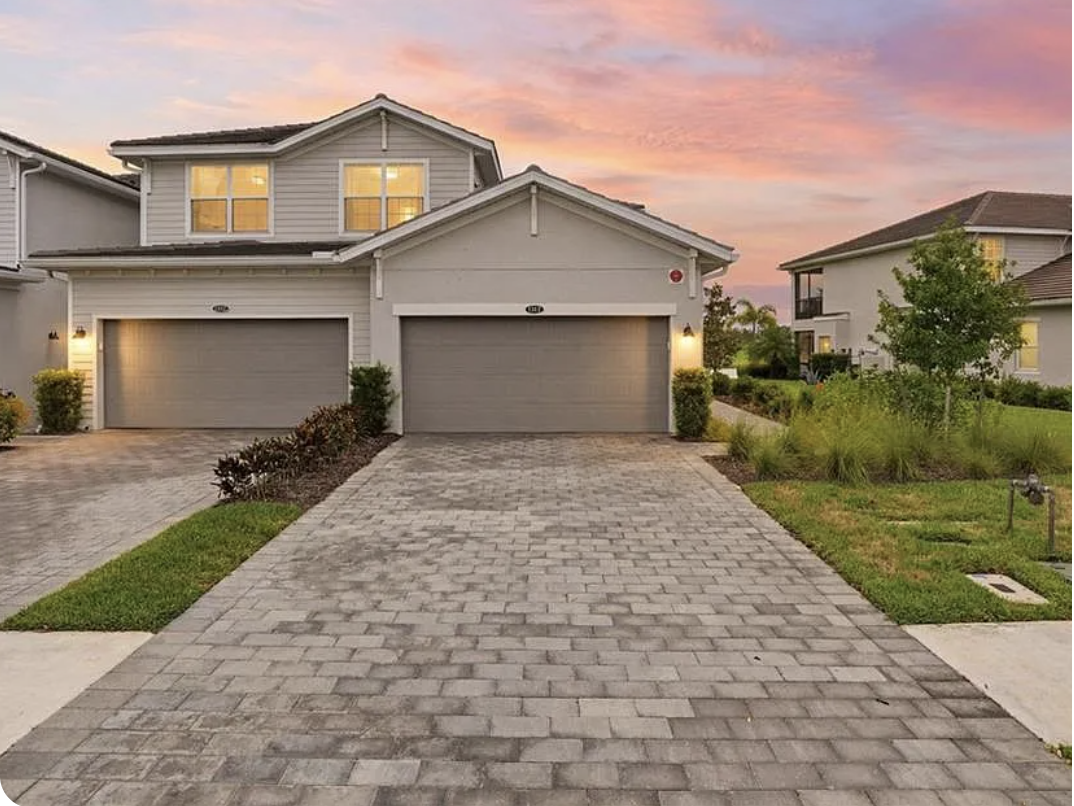 A spacious two-story rental home welcomes guests in a quiet residential neighborhood, featuring a paver driveway and attached two-car garage beneath a sunset sky.