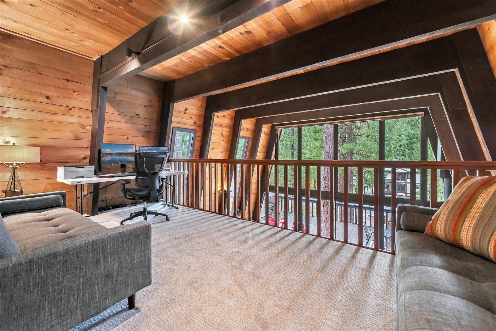 Cozy loft with striking wood beams, plush seating, and work desk overlooking towering pines through expansive windows.
