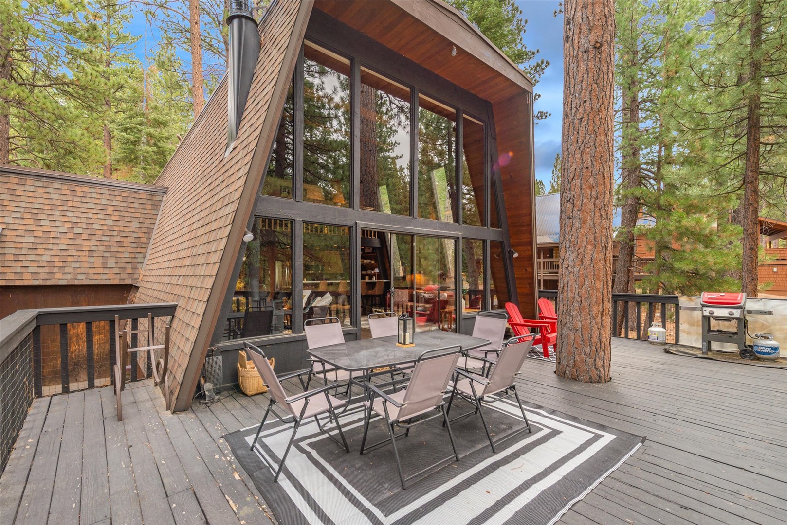 A classic A-frame cabin nestled among towering pines, featuring a spacious deck with dining space, mountain views, and a BBQ grill. The floor-to-ceiling windows frame the surrounding forest.