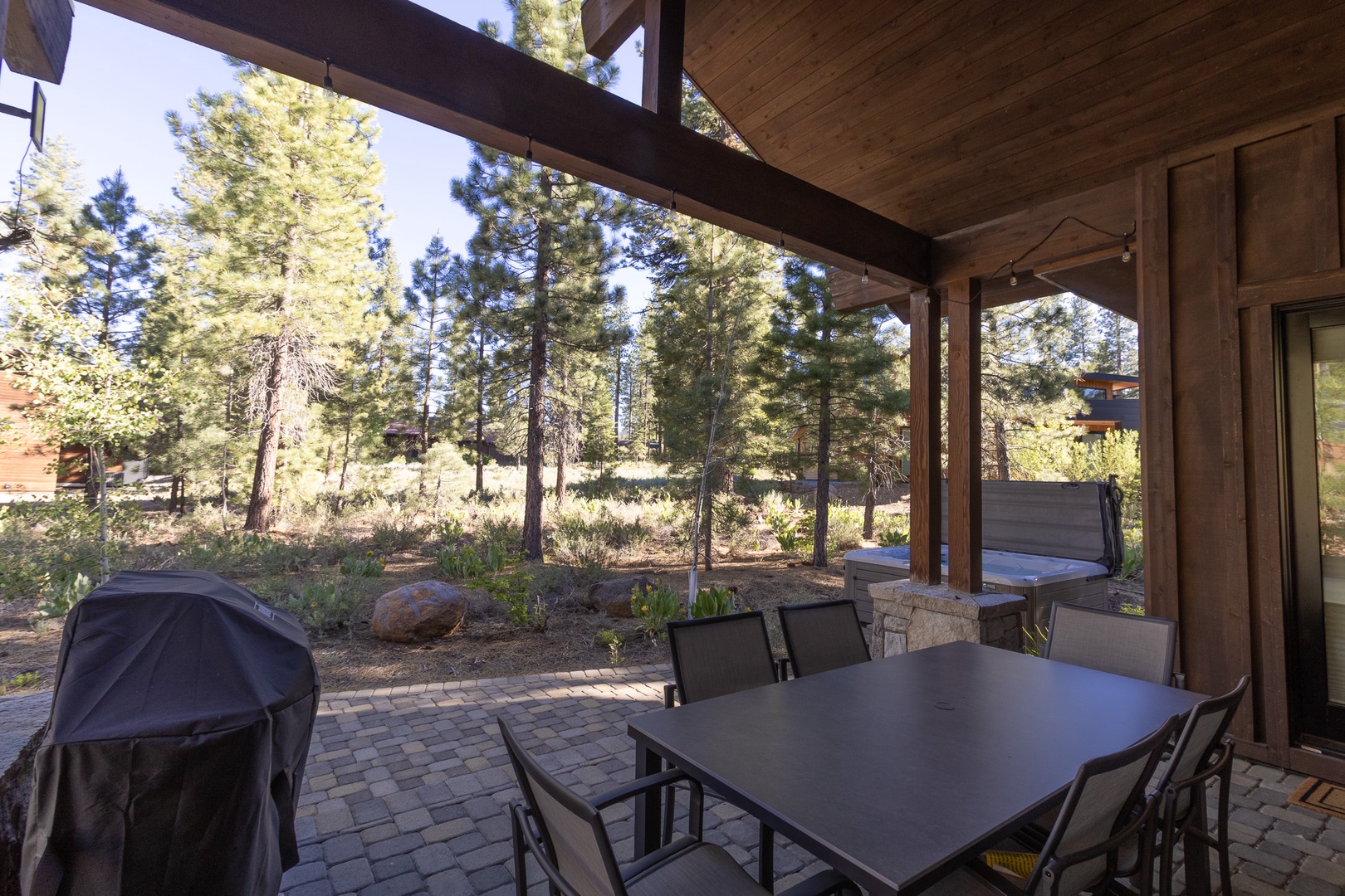 The covered patio overlooks a serene, forested landscape with towering pines framing the view. Brick pavers extend the living space outdoors, flanked by a grill for easy al fresco dining.