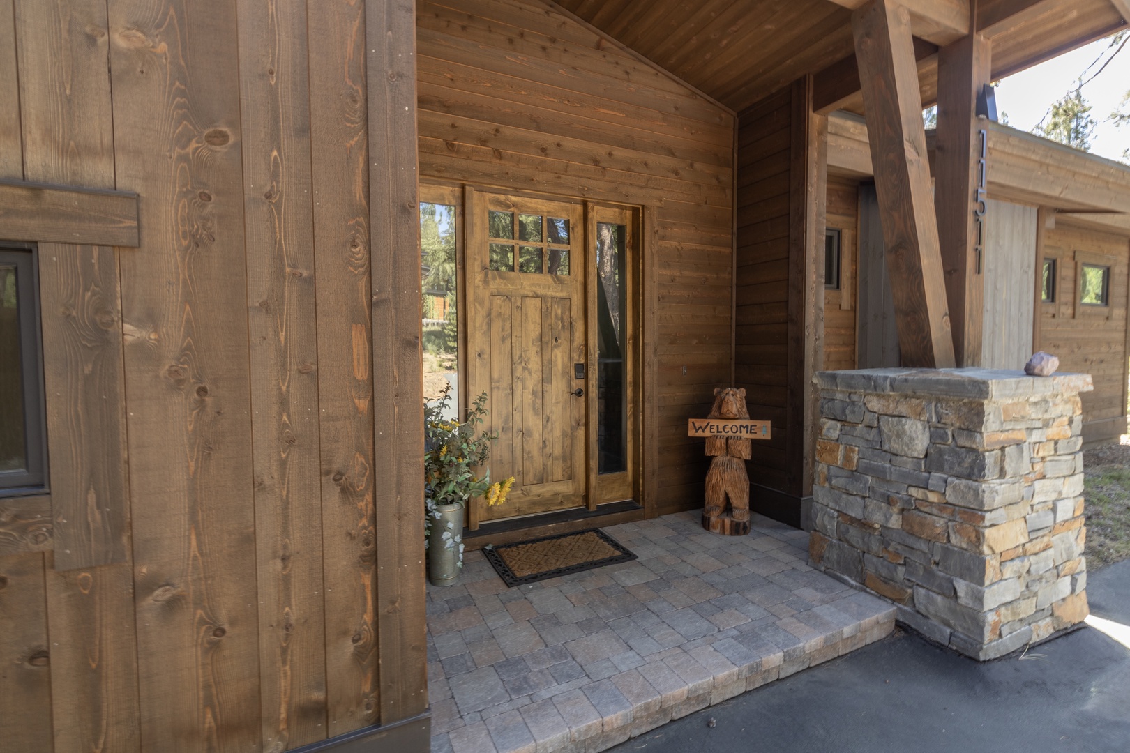 A welcoming covered entryway features natural wood and stone elements, with a charming bear statue greeting visitors beneath a protective overhang framed by timber beams.