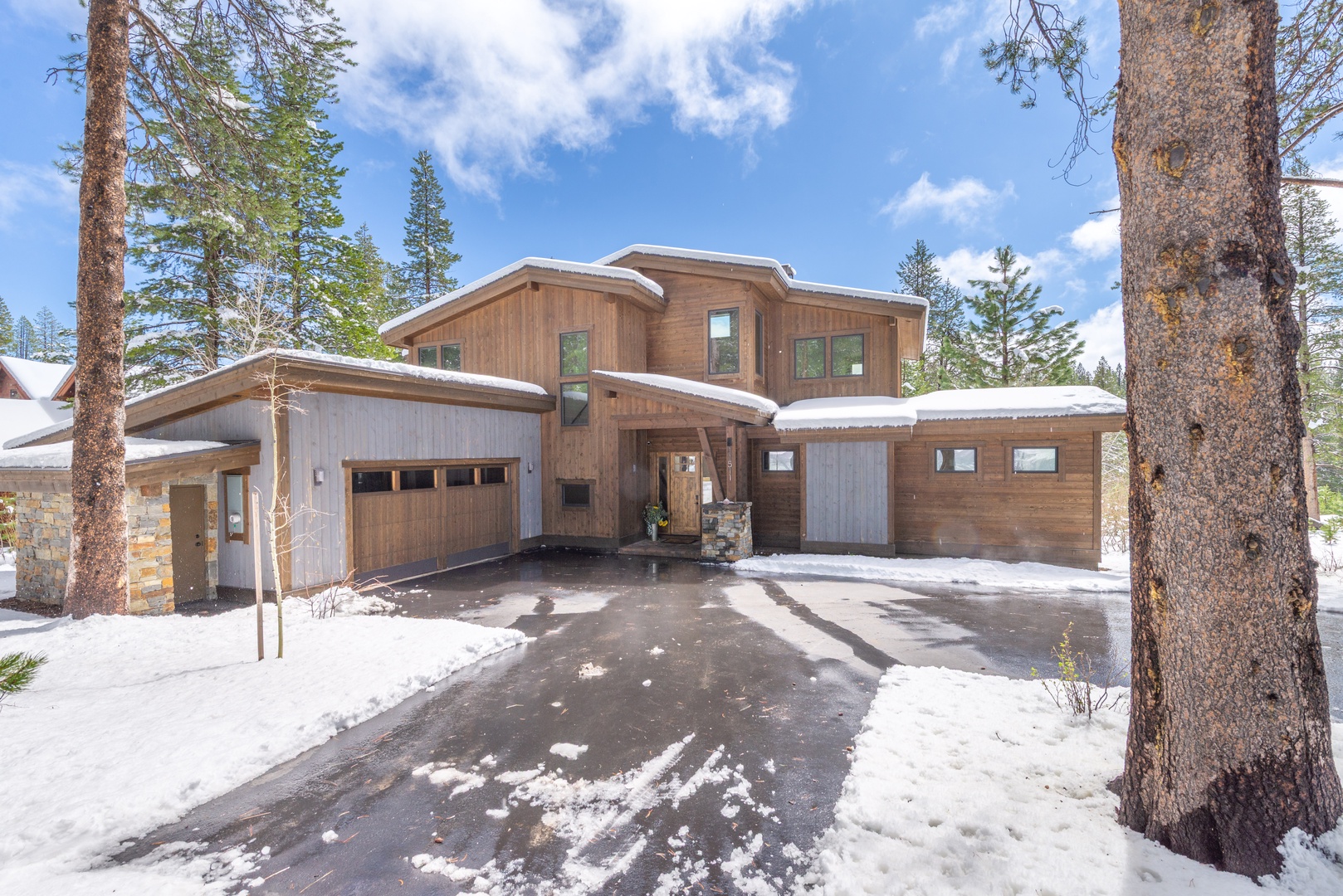 Modern mountain retreat with warm wood siding and stone accents nestled among towering pines in a snowy forest setting.